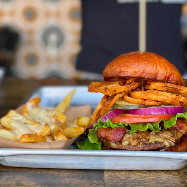 A Daiquiri Factory burger with bacon, onion rings, and fries on a metal tray.