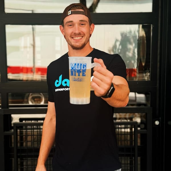 Smiling man in a Daiquiri Factory shirt holding up a "Mug Nite" beer mug.