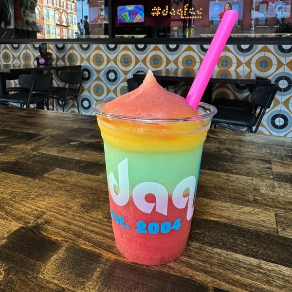 A colorful layered daiquiri with a pink straw sits on a wooden table inside Daiquiri Factory.