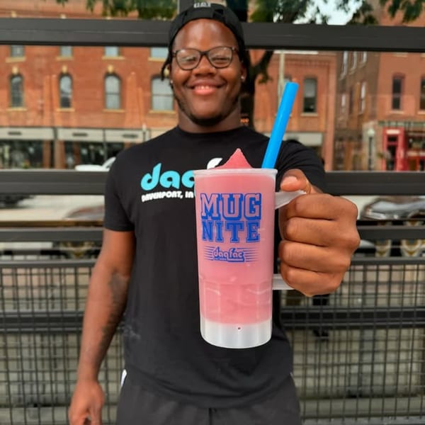 Smiling man in glasses and a black t-shirt holding out a pink Daiquiri Factory "Mug Nite" drink.