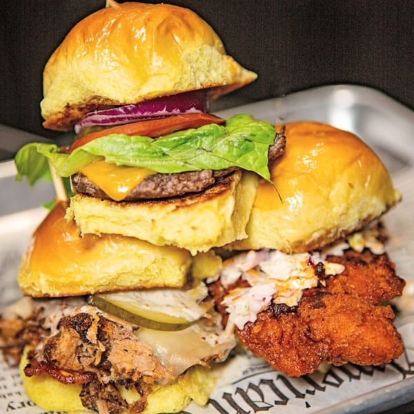 A trio of sliders: a cheeseburger, pulled pork, and fried chicken with coleslaw, on a metal tray.
