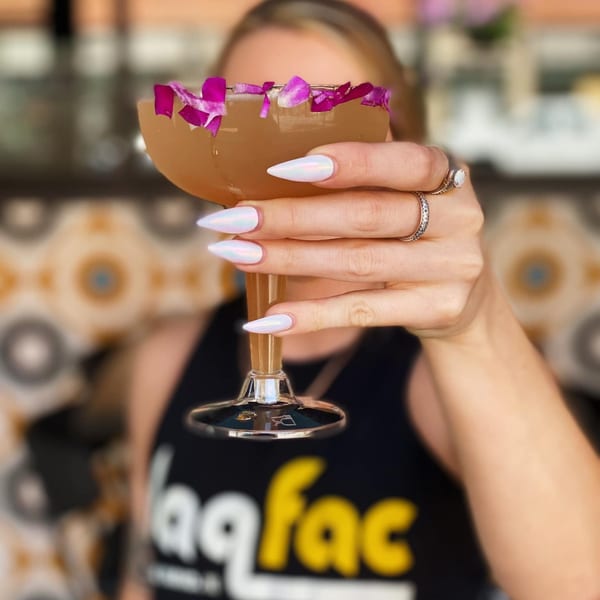 A woman in a Daiquiri Factory shirt holds a cocktail garnished with purple flower petals.
