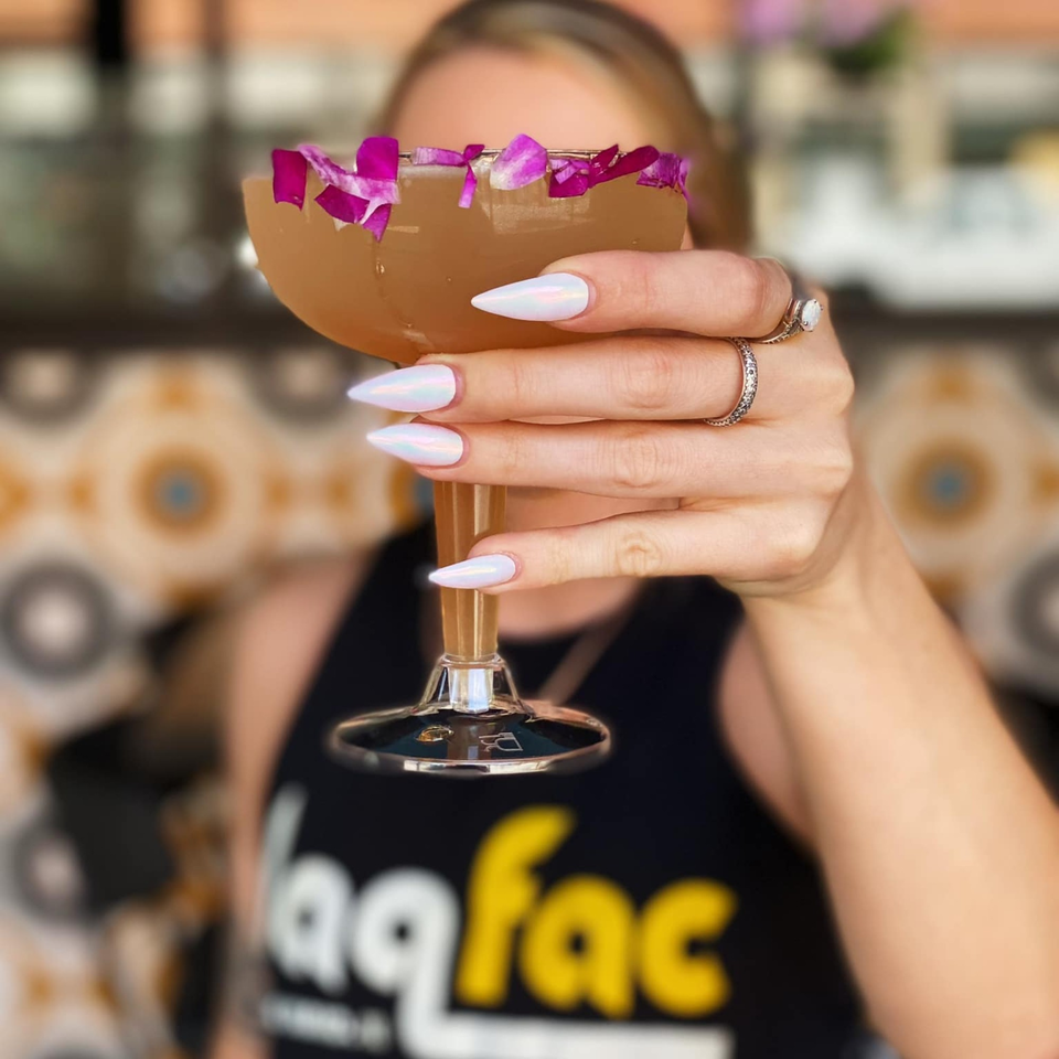 A woman in a Daiquiri Factory shirt holds a cocktail garnished with purple flower petals.