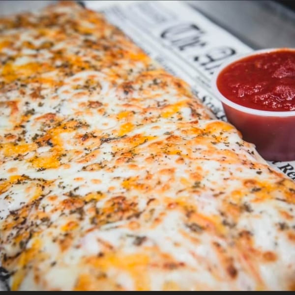 Cheesy breadsticks with herbs, served with a side of red marinara sauce on newspaper-patterned paper.