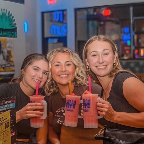 Three smiling women at Daiquiri Factory holding large pink frozen drinks with straws.
