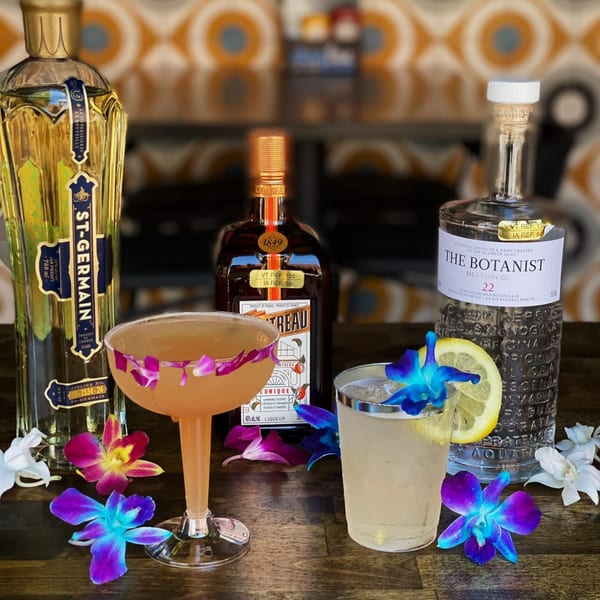 Two cocktails and three liquor bottles, garnished with colorful orchids, on a wooden surface.